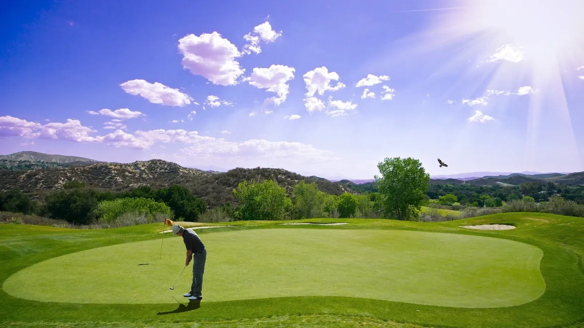 A golfer is putting on a beautifully manicured green, with distant hills and a partly cloudy sky setting a scenic backdrop. The lush landscape and open space evoke the atmosphere of a country club setting, even though club amenities are not included.