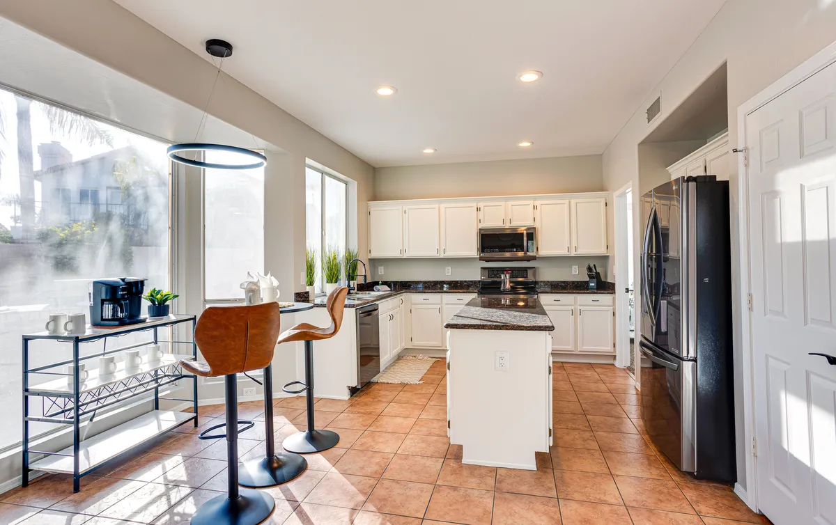 A bright, modern kitchen with a spacious island, bar stools for casual seating, large windows flooding the space with natural light, and premium appliances lining the walls. The cheerful, relaxed mood invites both cooking and socializing.