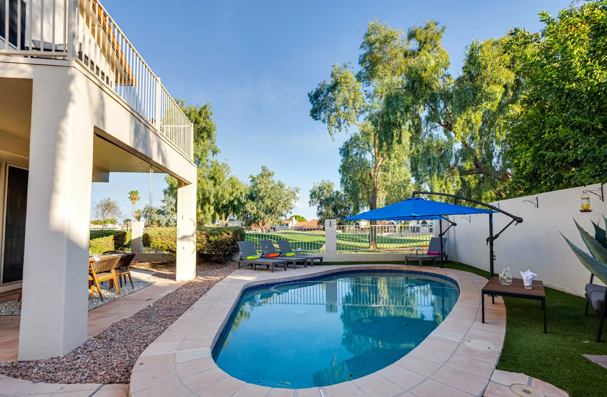 A tranquil backyard scene featuring a sparkling swimming pool surrounded by plush lounge chairs, an outdoor umbrella, and a stylish table with decorative accents. Lush landscaping adds to the serene atmosphere, with the setting sun casting a warm glow across the patio.
