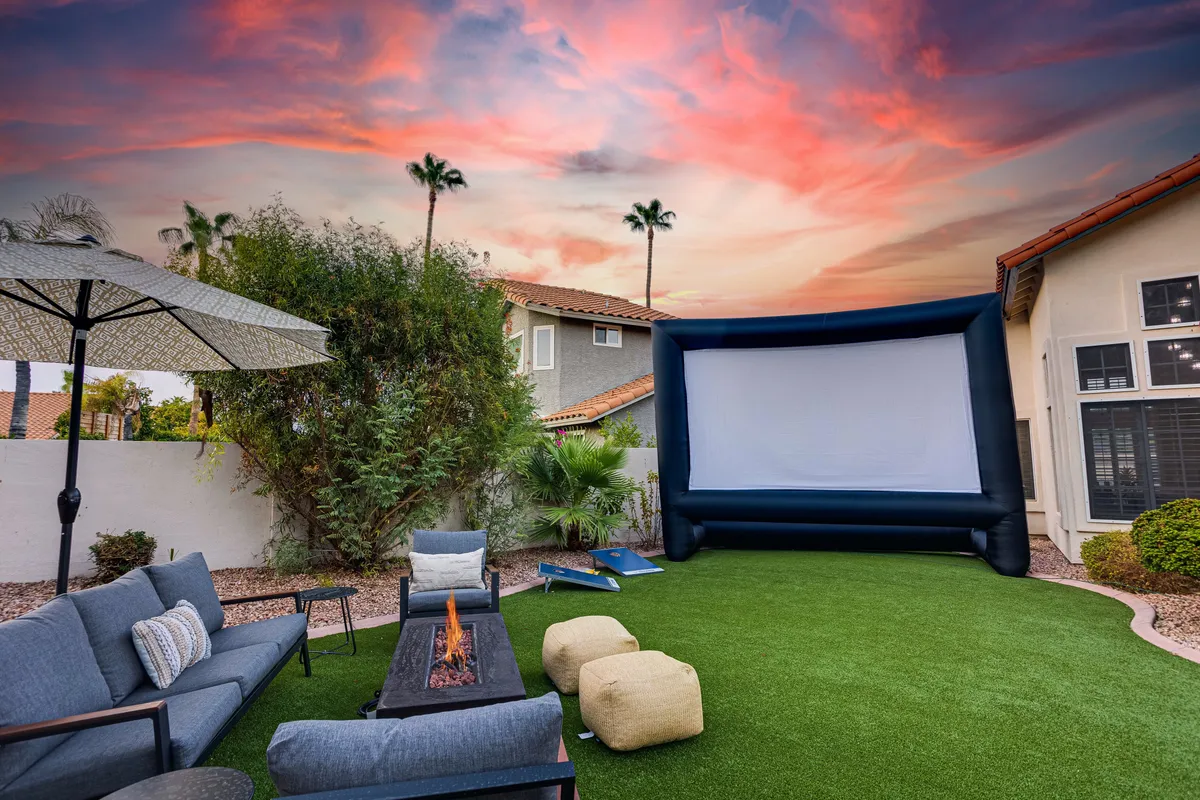 An inviting backyard set up for outdoor movie nights, featuring an inflatable movie screen, a cozy fire pit, comfortable seating, and surrounded by palm trees. The laid-back setting is perfect for group gatherings after a day of sports or golf.