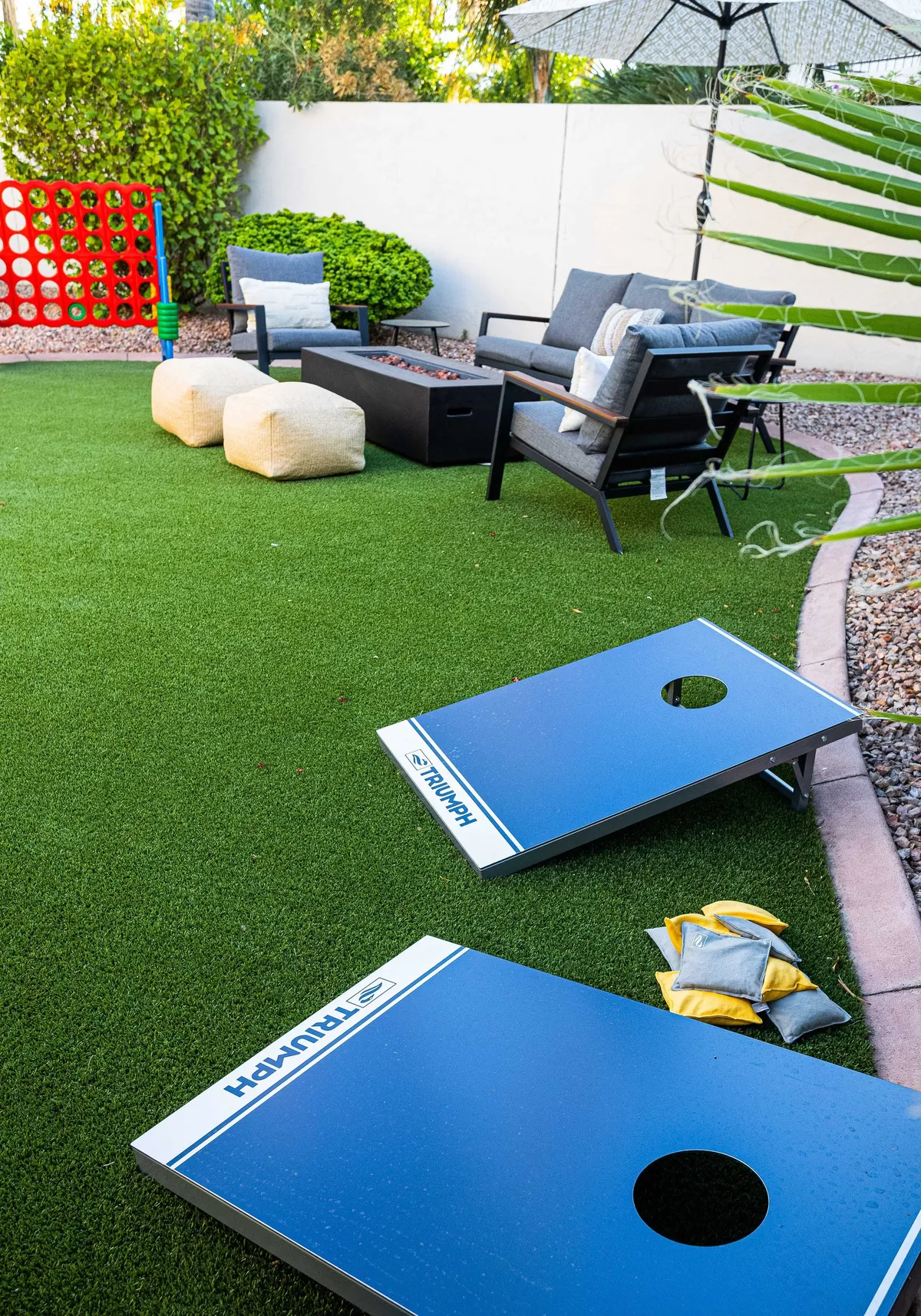 The photo shows an exterior backyard scene with a lawn game (cornhole) set up, a comfortable seating area, and decorative plants. The ambiance is playful and relaxed, ideal for socializing and outdoor family activities.