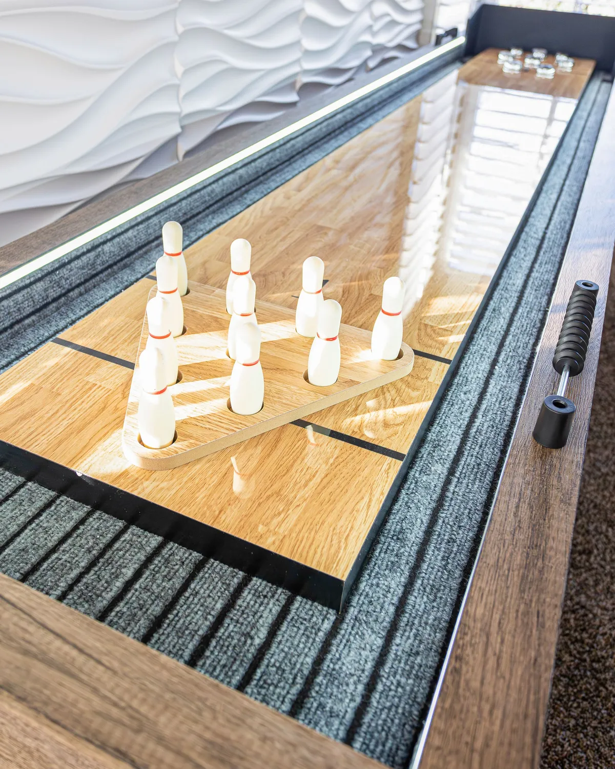A vibrant game room scene with a shuffleboard table, bowling pins, a polished wooden surface, and a decorative wall. The space is designed for playful interaction and informal entertainment.
