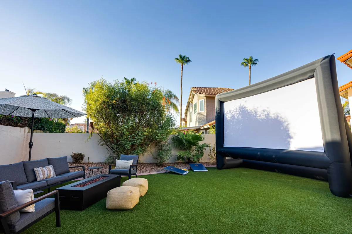 An inviting patio area featuring a large inflatable outdoor movie screen, cozy fire pit, comfortable seating, and swaying palm trees overhead. The setting is casual and festive, embodying the excitement of an open-air cinema experience under the Arizona sky.