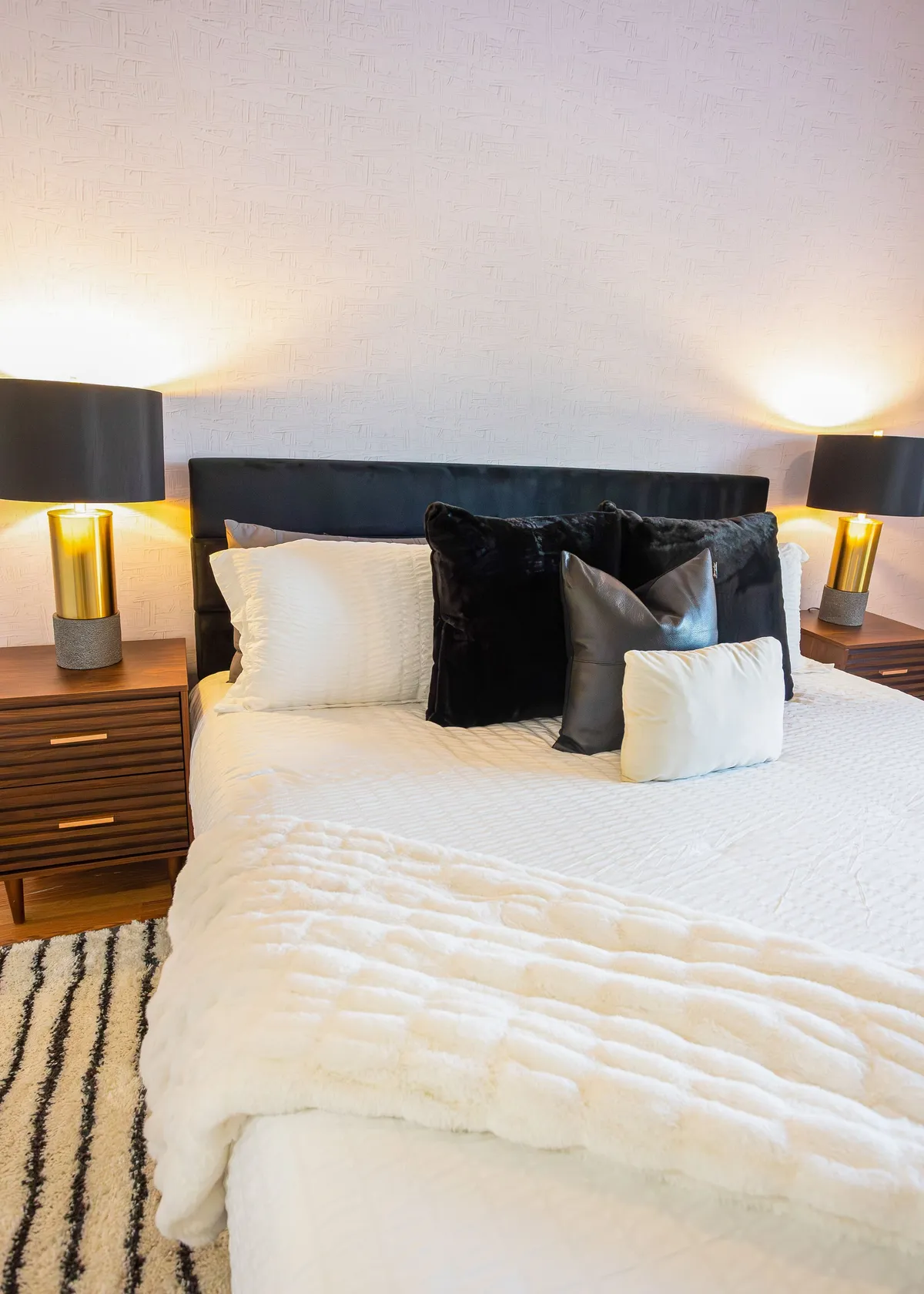 A calm, softly lit queen bedroom with a cozy bed covered in decorative pillows, flanked by nightstands and reading lamps. The neutral tones and simple décor evoke a sense of peace and restful retreat.