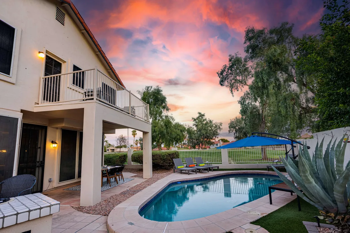 A serene view of a sparkling swimming pool bordered by lush greenery, with inviting patio seating and a balcony overhead. The sky is painted in soft sunset colors, enhancing the tranquil, inviting atmosphere. The chic deck furniture is positioned for both sunbathing and relaxed conversation, with easy access to the pool.