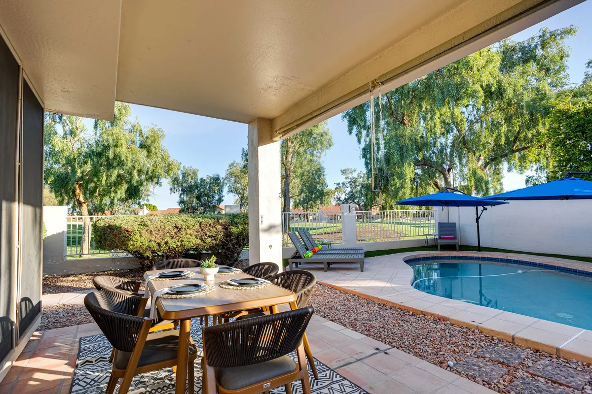 A relaxing outdoor scene with a swimming pool, lounge chairs, umbrellas, a dining table, and lush green surroundings. The seating is set up for both sunbathing and outdoor dining, inviting both relaxation and social gatherings beside the water.