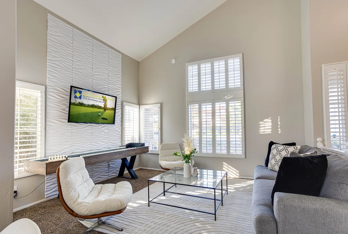 A contemporary living room featuring a large TV mounted on a textured wall panel, a glass coffee table, stylish armchairs, and a decorative plant. The space is designed for comfort and entertainment, encouraging relaxation and social gatherings.