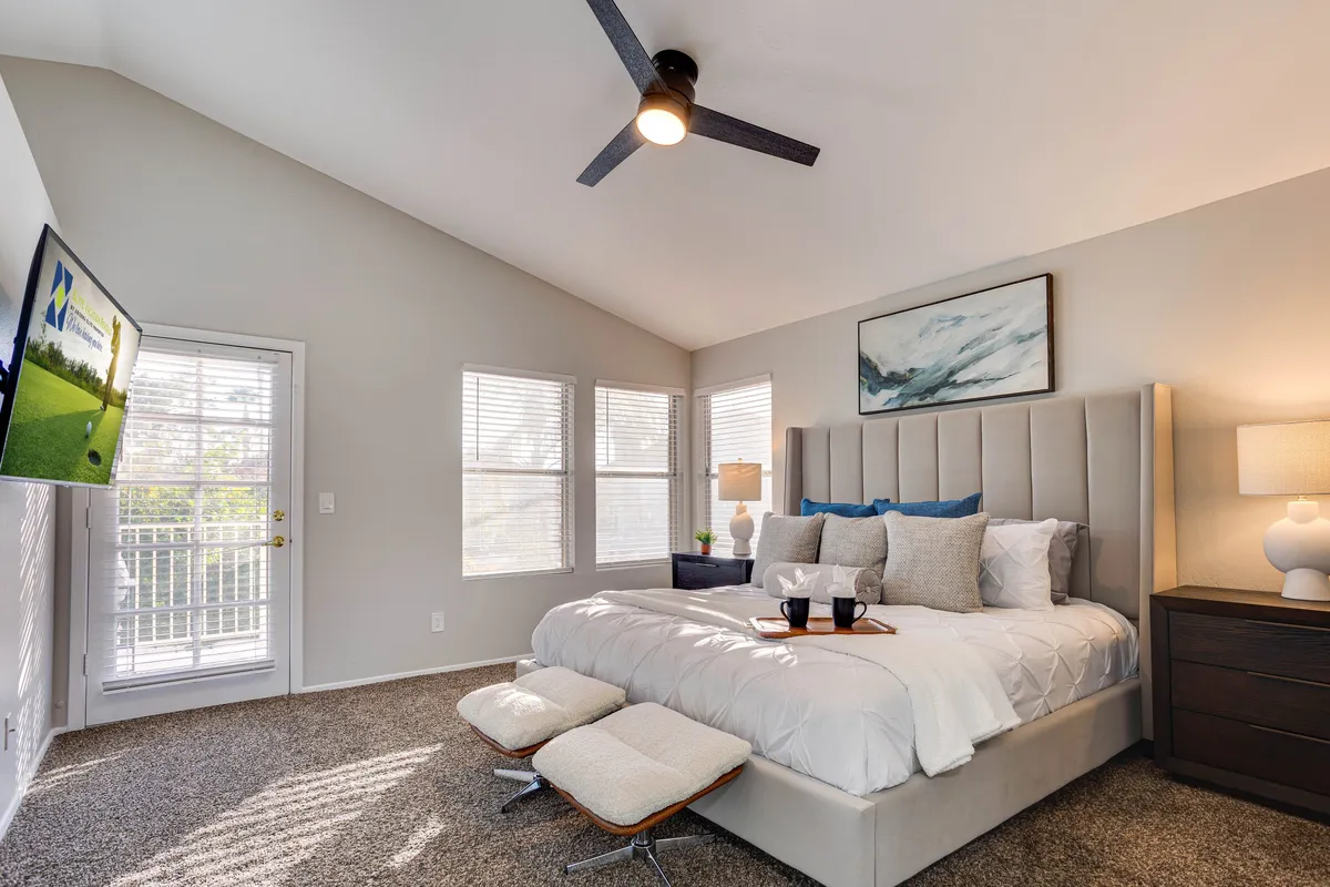 A spacious king-sized bedroom featuring a large, inviting bed with crisp bedding, nightstands with lamps, a TV mounted on the wall, and expansive windows allowing abundant natural light to fill the space. The serene palette and uncluttered décor create an atmosphere of tranquility and comfort.