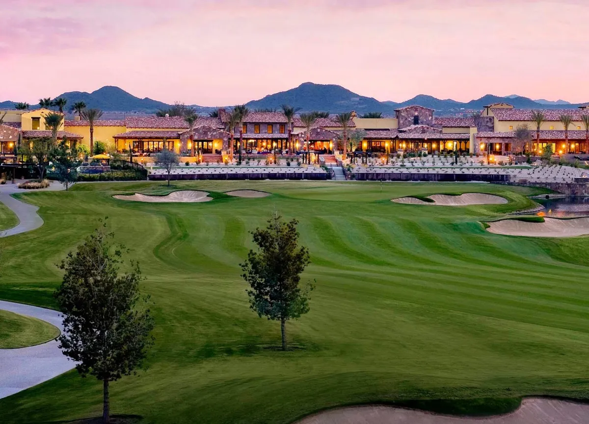 A breathtaking view of a championship golf course with a clubhouse in the background, framed by palm trees and dramatic mountain scenery. The golden hour lighting and lush fairways emphasize the upscale, resort-like surroundings nearby.